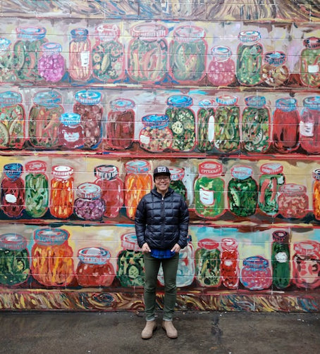 owner Asano Otsu stands in front of a mural of painted jars of pickles